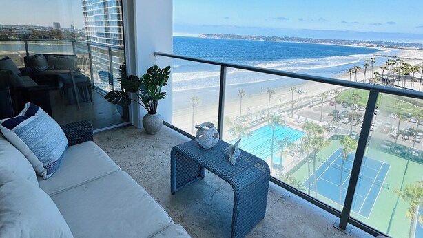 Een balkon met uitzicht op de oceaan en een tennisbaan. Er is een bank en een klein tafeltje met een decoratief object erop.