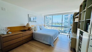 Een moderne slaapkamer met een groot raam, een bed, een dressoir en een inbouwkast.