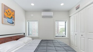 A bedroom with a bed, white walls, a painting of flowers, two windows with blinds, an air conditioner, and a wardrobe. - @StayatNoMA (Washington)