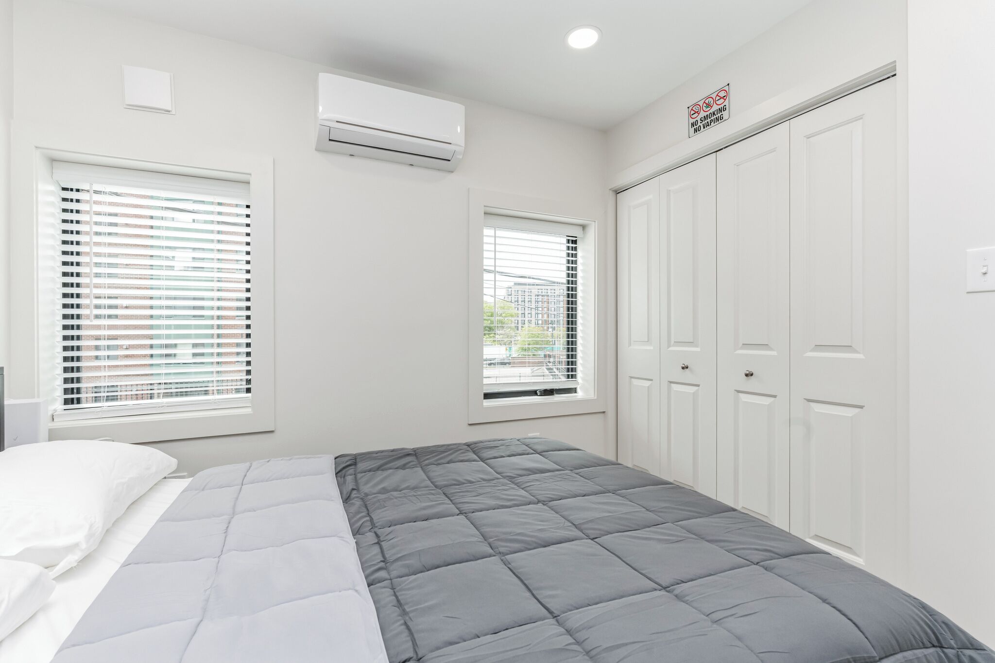 Een slaapkamer met een bed, twee ramen met luxaflex, een airconditioner en een kledingkast.