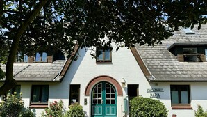 A white building with a green door, a sign reading "FRIESENHAUS MARIA," and a small garden area with flowers and shrubs.