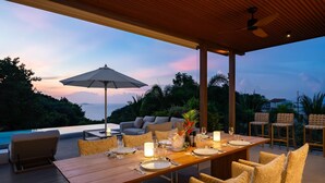 An outdoor dining area with a wooden table set for four, surrounded by cushioned chairs. There is a large umbrella providing shade, and the setting overlooks a body of water with a view of the horizon. - Villa Divano by Sublime Samui Villas (Koh Samui)