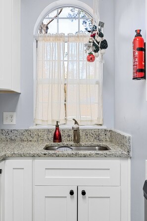 Cozinha com bancada de granito, armários brancos, pia de aço inoxidável, torneira vermelha e uma janela com cortinas leves.