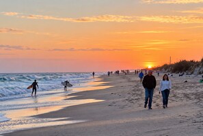 Escena de playa con gente caminando por la orilla, un atardecer al fondo y olas rompiendo en la playa.