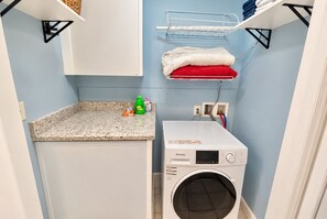 A compact laundry room with a washing machine, a countertop with cleaning supplies, and a shelf with folded towels. - Top-Rated ILM Oasis: Spacious, Pet-Friendly, Pool! (Wilmington)