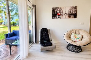 A room with a glass door leading to a patio, a blue sofa, a black baby stroller, a beige chair with a floral cushion, and a wall-mounted painting of a cityscape. - Top-Rated ILM Oasis: Spacious, Pet-Friendly, Pool! (Wilmington)