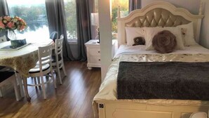 Ein Schlafzimmer mit einem Bett, einem Nachttisch, einem Stuhl, einem kleinen Tisch mit Blumen und Blick ins Freie durch ein Fenster.