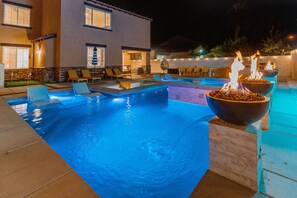Zona de piscina exterior bien iluminada, con sillones de descanso y una chimenea.
