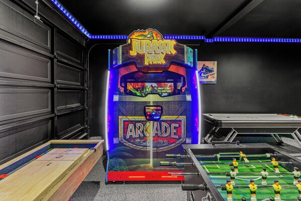 A room with a foosball table, a pool table, and a sign for 'Jurassic Park Arcade'.
