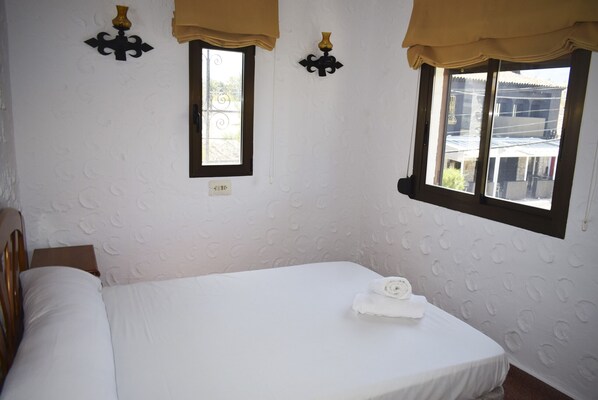 Una cama bien hecha con una toalla enrollada, una ventana con cortina y apliques de pared.