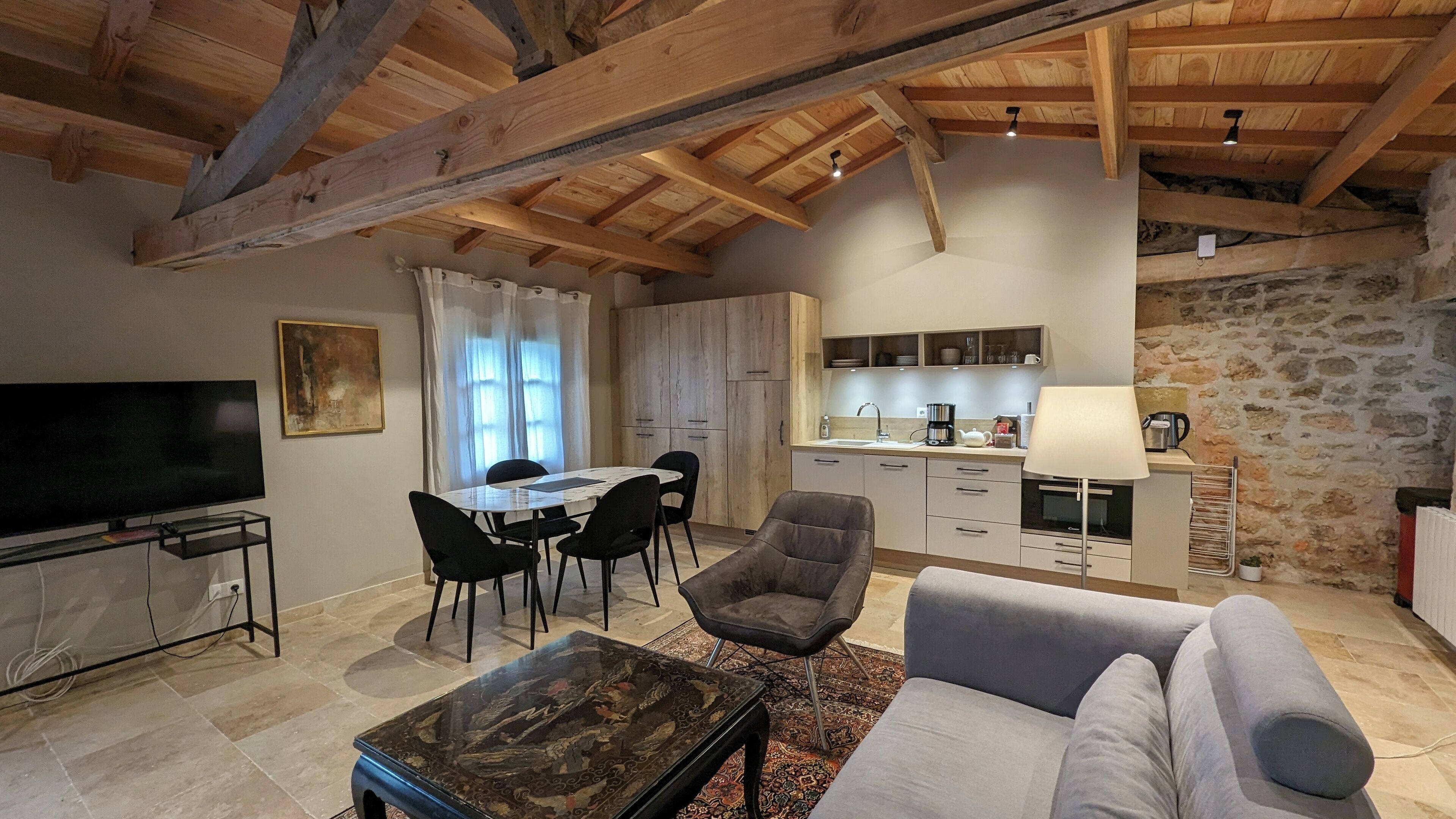 Un salon spacieux doté d’un téléviseur à écran plat, un coin repas avec une table et des chaises, une cuisine équipée d’appareils électroménagers modernes et un mur en pierre.