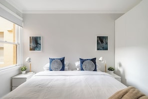 A modern bedroom with a large bed, two bedside tables, a window, and two wall-mounted paintings. - Renovated Harbourfront Home Views of Harbour/Pool (Manly)