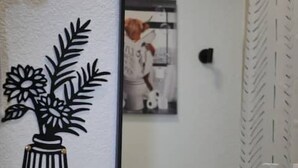 Ein Badezimmer mit einem weißen Waschbecken, einem schwarzen Wasserhahn und einem Spiegel, der eine Person reflektiert. Darüber befindet sich eine schwarz-weiße Wanddekoration.
