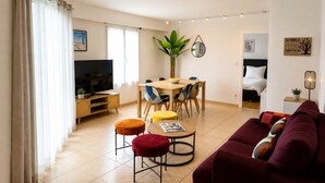 A living room with a sofa, ottomans, a dining table with chairs, a TV, and a plant.