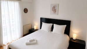 A neatly made bed with white linens, two bedside tables with lamps, a framed artwork on the wall, and a window with sheer curtains.