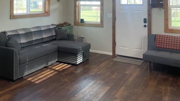 A spacious living room with a sectional sofa, a door, and two windows with blinds. - Tiny house hideaway just outside of town (Cody)