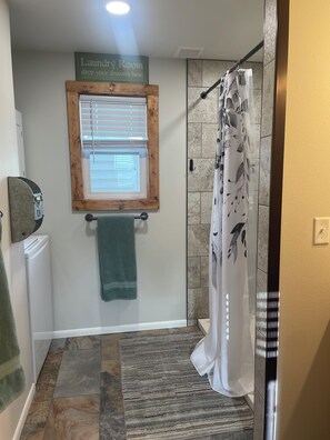 Une salle de bain avec une douche, un porte-serviettes, un panneau indiquant qu’il s’agit d’une buanderie, et une fenêtre avec des stores.