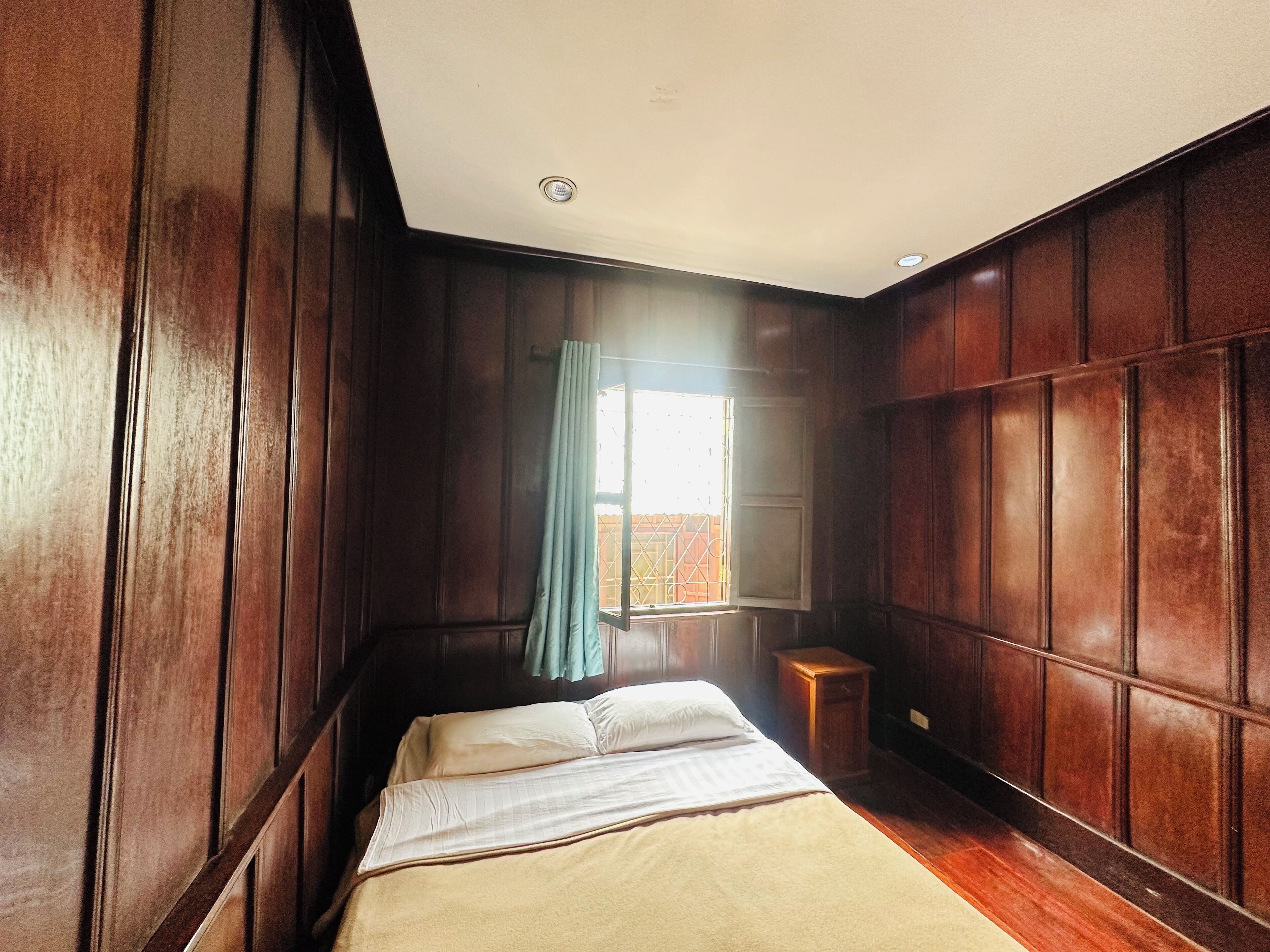 Ein Zimmer mit Holzwänden, einem Bett mit weißen Bettwäsche, einem Fenster mit Vorhang und einem kleinen Nachttisch aus Holz.