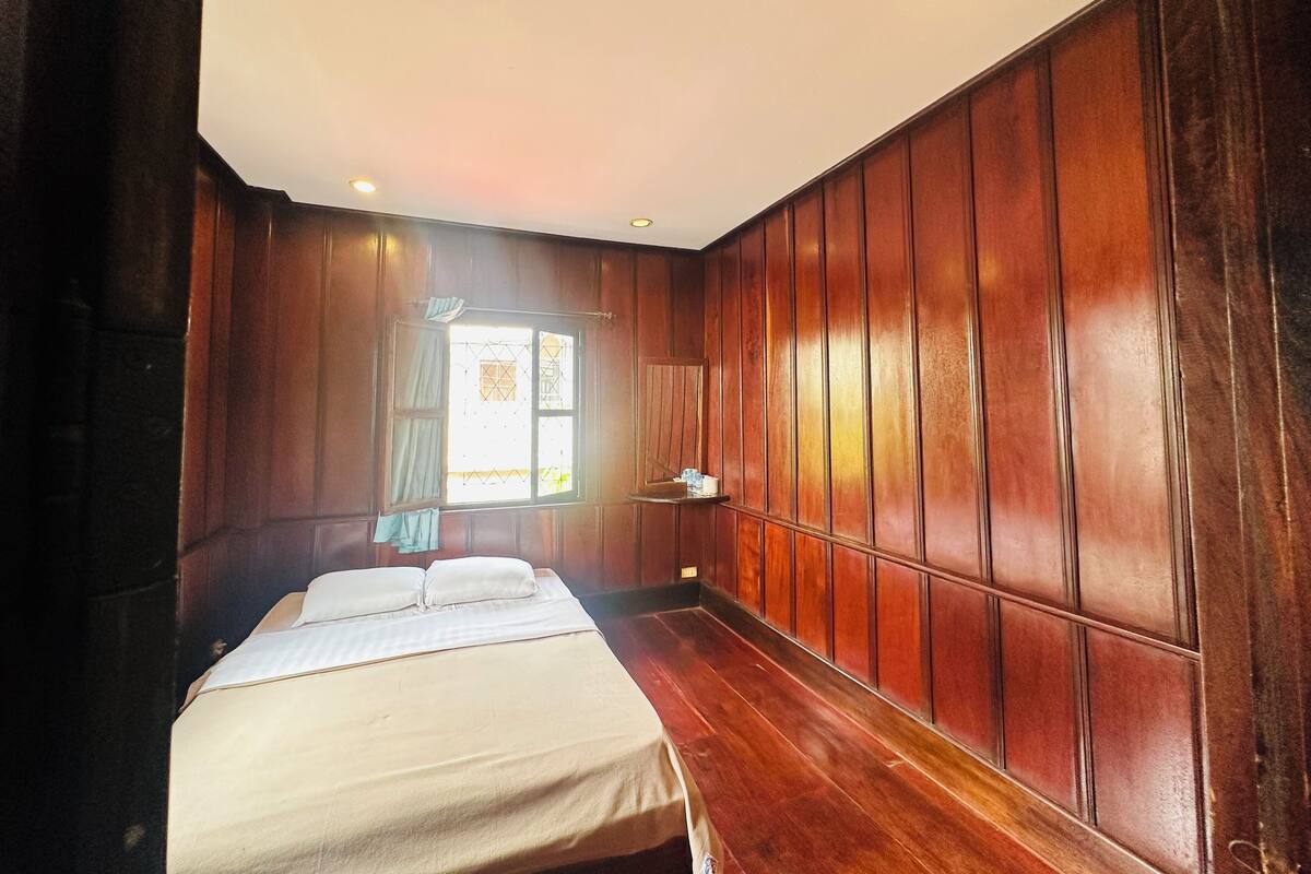 A room with wooden paneling, a bed with white bedding, a window with curtains, and a small shelf with items.