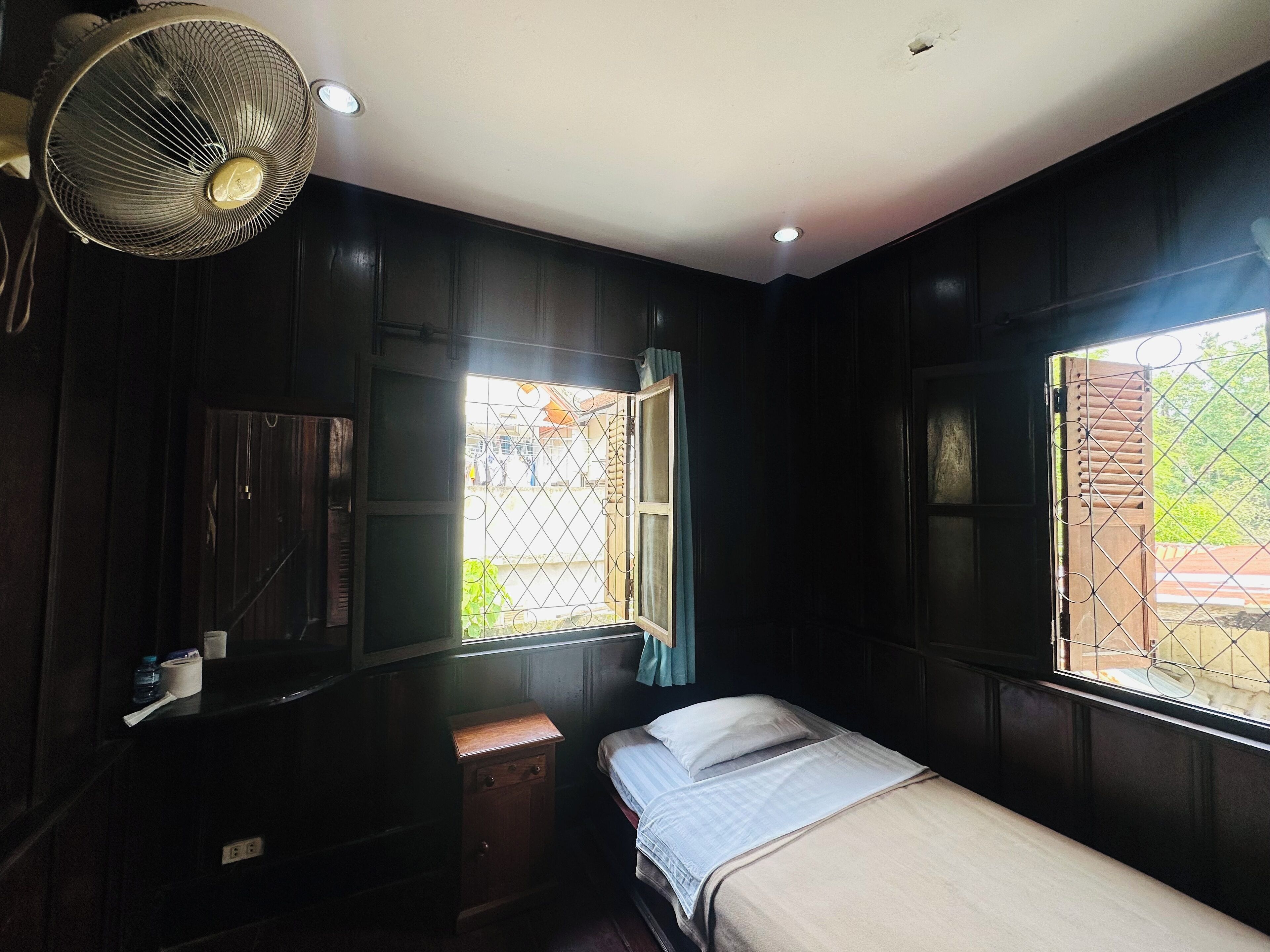 A small, dimly lit room with a single bed, a wooden cabinet, and a window with a metal grill.