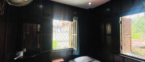 A small, dimly lit room with a single bed, a wooden cabinet, and a window with a metal grill.