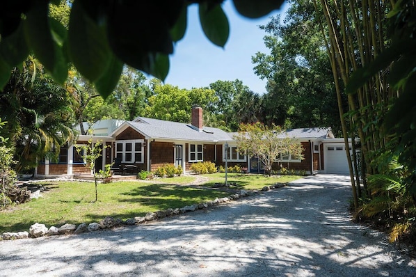 Traditional two-story house with a brick facade, surrounded by lush greenery and a gravel driveway.