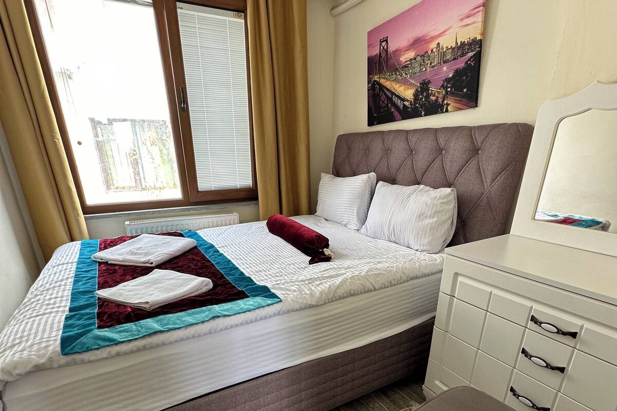 A hotel room with a bed, a bedside table, a chair, a radiator, and a window with curtains.