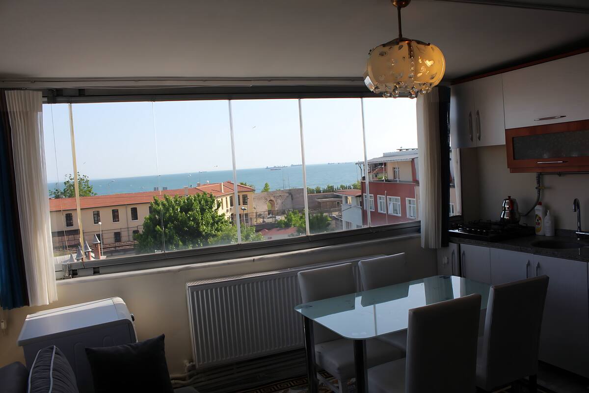 A room with a large window, a table, chairs, a radiator, and a kitchen area with a sink and cabinets.