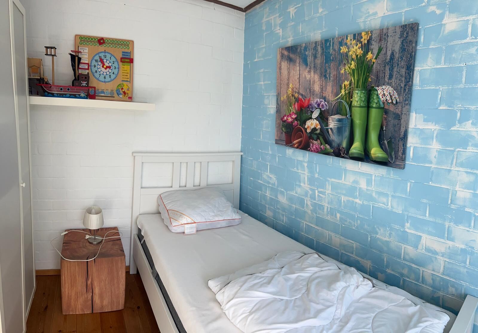 A small, neatly arranged bedroom with a single bed, a wooden nightstand, a shelf with a clock, and a wall-mounted painting of flowers and gardening tools.