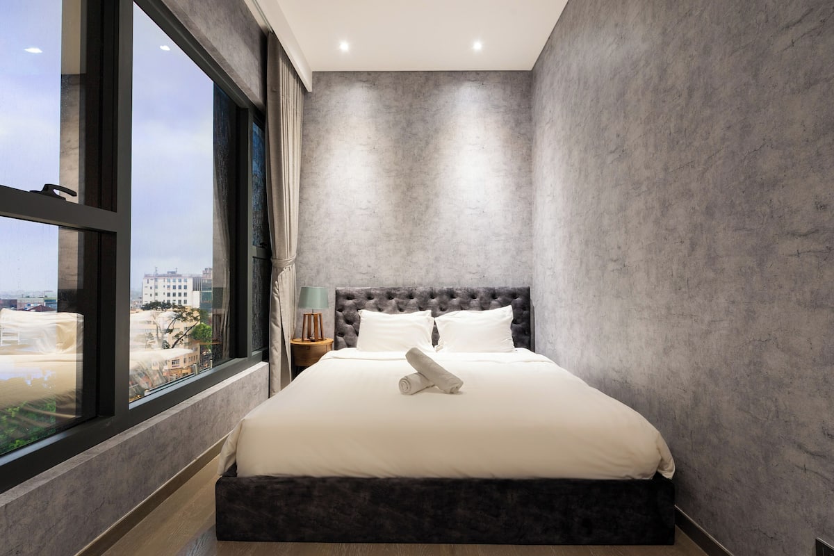 Une chambre moderne avec un grand lit, une vue sur la ville et un mur gris à texture.