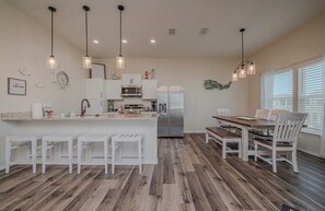 A modern kitchen with white cabinetry, stainless steel appliances, and a wooden dining area with white chairs. - Coastal Getaway 3BR 2BA, sleeps up to 8, w pool, balcony, mins to beach & marina (Rockport)