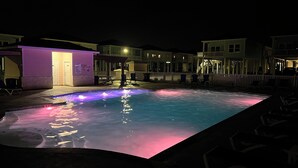 Una piscina amb un element aquàtic de nit, envoltada d'edificis amb una tanca blanca i zones d'estar a l'aire lliure.