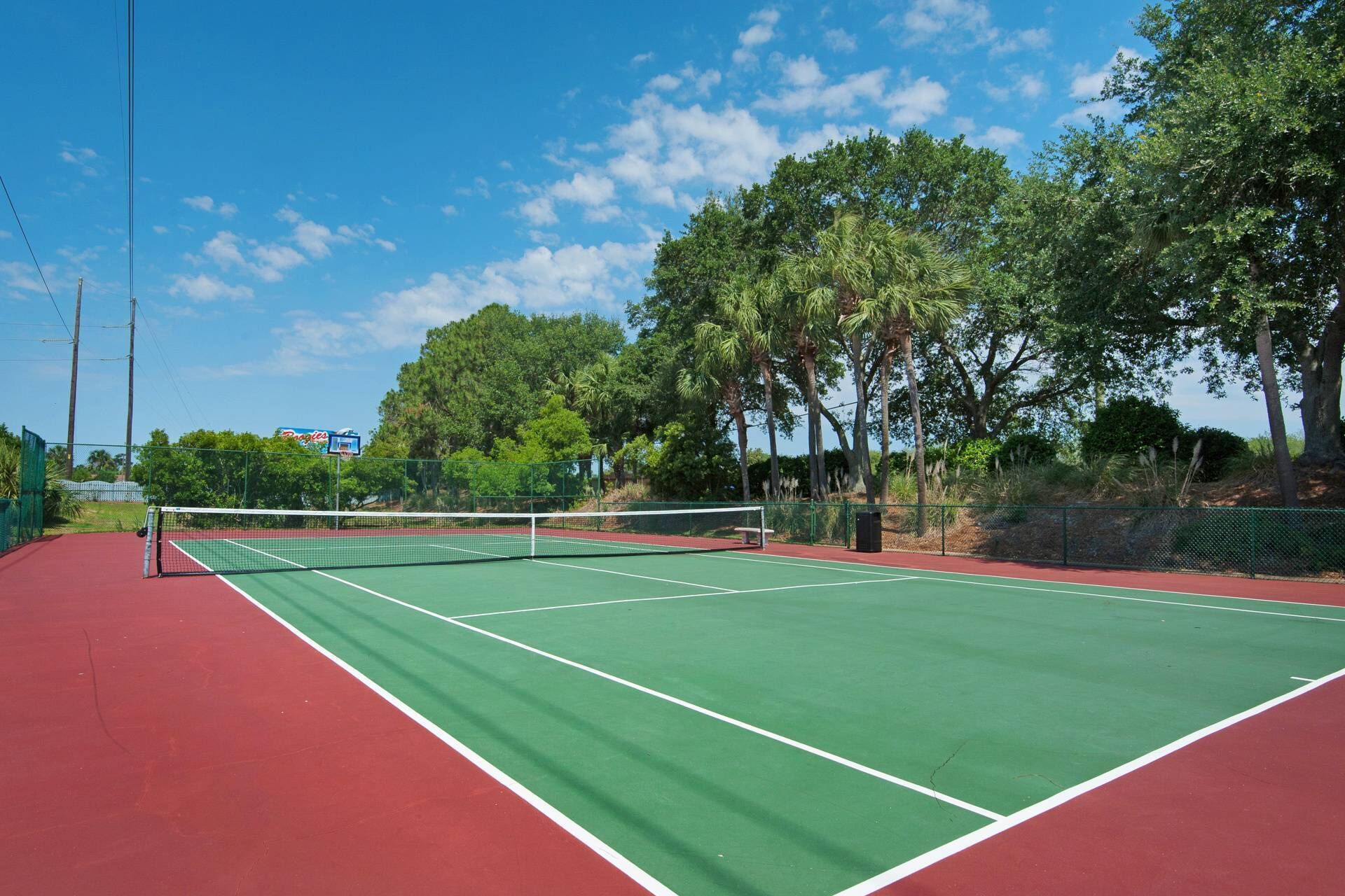 An outdoor tennis court with a green playing surface and red borders, surrounded by trees and a clear sky.
