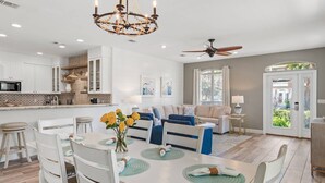 A modern kitchen with white cabinets, a dining area with a white table and blue chairs, a living room with a beige sofa, and a ceiling fan. - Pet & Family Friendly, Walk or Tram 2 Private Beach Access Pavilion, Pools, Tennis - Emerald Shores (Miramar Beach)