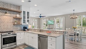 A modern kitchen with white cabinetry, a stainless steel stove, and a dishwasher. There is a dining area with a table and chairs, and a TV mounted above the counter. - Pet & Family Friendly, Walk or Tram 2 Private Beach Access Pavilion, Pools, Tennis - Emerald Shores (Miramar Beach)
