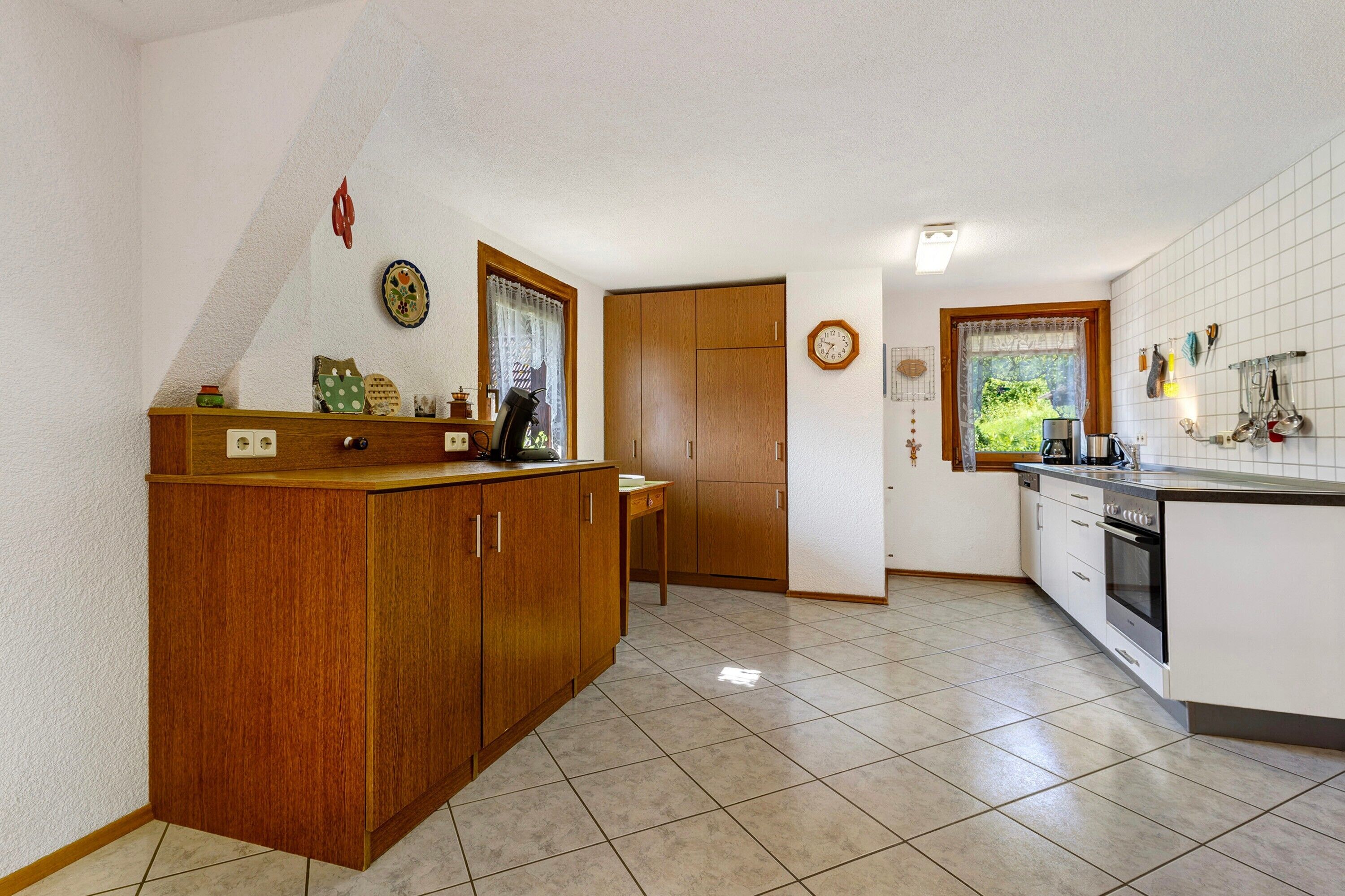 Una cocina con armarios de madera, suelo de baldosas, una ventana con cortinas y un reloj en la pared.