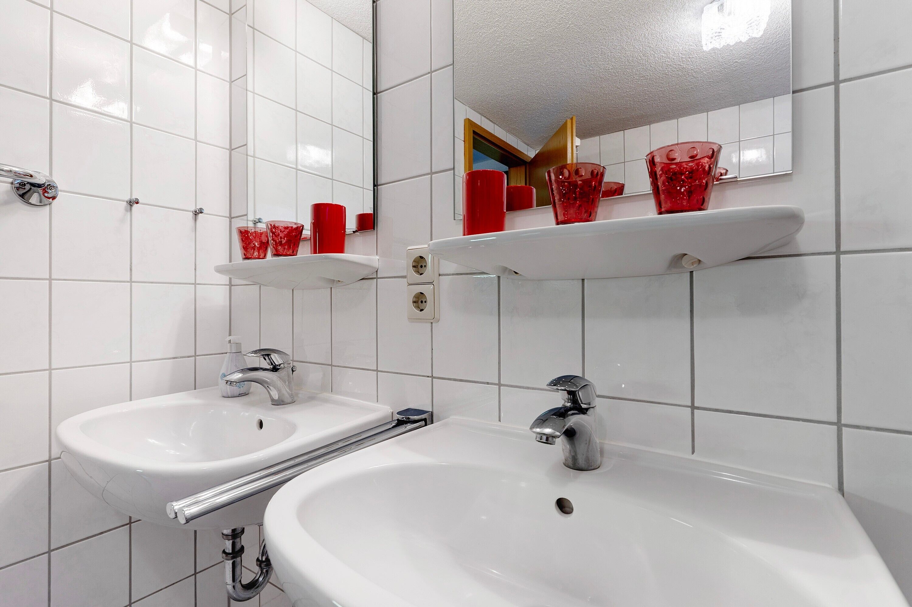 Un baño con dos lavamanos, un espejo y vasos de vidrio rojo en la repisa.