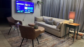 A modern living room with a flat-screen TV mounted on the wall, a sofa, a coffee table, and a lamp on a side table. - Newly renovated home near Hard Rock Stadium and major highways! (Miami Gardens)