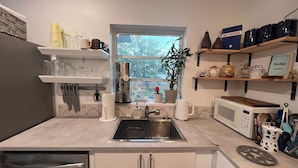 A modern kitchen with a stainless steel sink, a coffee maker, and a microwave. There are shelves with kitchenware and a window with a view of trees. - Newly renovated home near Hard Rock Stadium and major highways! (Miami Gardens)