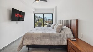 Ein modernes Schlafzimmer mit einem großen Bett, einem an der Wand montierten Fernseher, einem Deckenventilator und Blick auf die Stadt durch ein großes Fenster.