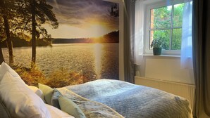 Ein Schlafzimmer mit Bett, Kissen, einem Fenster mit Blick auf Grünflächen und einem Wandbild, das einen Sonnenuntergang über Wasser zeigt.