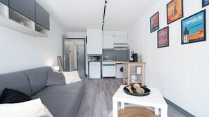 A compact living space with a grey sofa, a small white table, a kitchenette, and a door leading to another room.