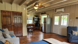 A cozy living room with a sofa, a blue rug, and a chair. A kitchen with a refrigerator, stove, and microwave is visible in the background. - Tack Room @ Redemption Ranch: Grill, Deck, Peaceful, Unique, Comfortable (Marionville)