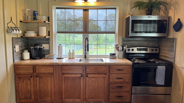 A kitchen with wooden cabinets, a stainless steel stove, a microwave, and a window with a view of greenery. - Tack Room @ Redemption Ranch: Grill, Deck, Peaceful, Unique, Comfortable (Marionville)