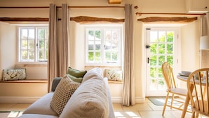Ein gemütliches Zimmer mit einer Couch, einem Holzstuhl und einem Esstisch. Es gibt Fenster mit Vorhängen, eine Tür und einen Blick auf Grünfläche.