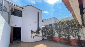 A courtyard with a tiled floor, a white building with a red and white wall, and a metal fence. - House Near The Santiago River (San Luis Potosi)