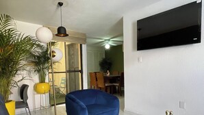 A modern living room with a blue armchair, a grey sofa, a flat-screen TV mounted on the wall, a laptop on a wooden table, and a dining area in the background. - House Near The Santiago River (San Luis Potosi)