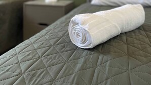 A neatly made bed with a rolled white towel on a green quilted bedspread, a tufted headboard, and a bedside table with a lamp. - House Near The Santiago River (San Luis Potosi)