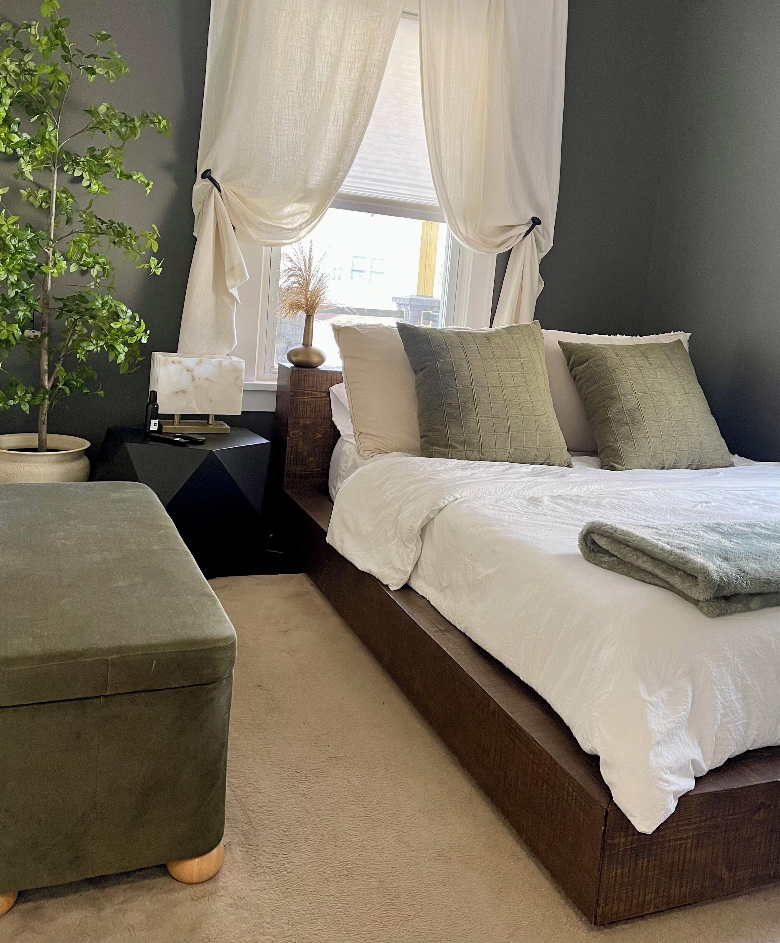 Een slaapkamer met een bed, nachtkastje, lamp, een bloempot en een raam met gordijnen.