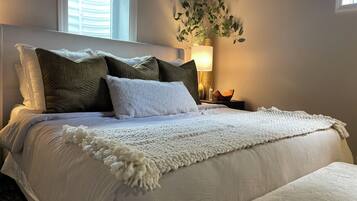 A neatly made bed with white bedding and a fluffy white throw, a bedside lamp, and a plant on a nightstand.